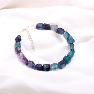 May include: A beaded bracelet featuring rectangular fluorite stones in shades of purple, teal, and blue. The bracelet has a silver-colored chain clasp. The stones are faceted, catching the light.