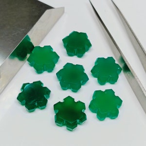 May include: Seven green snowflake-shaped cabochons, each with six points, are arranged on a white surface. The cabochons are smooth and polished, with a slight sheen.