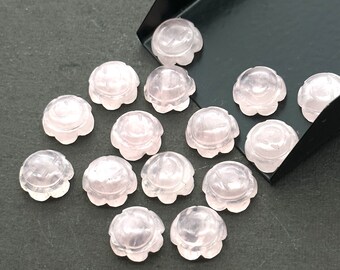 Puede incluir: Una colección de 14 cabujones de cuarzo rosa pálido, cada uno tallado con un diseño de flor. Los cabujones están dispuestos sobre una superficie negra.