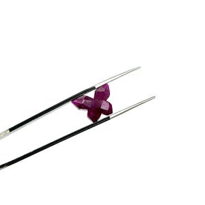 May include: A deep red, faceted gemstone in the shape of a butterfly is held by tweezers. The butterfly gem has multiple facets that reflect light. The tweezers are silver. The gem is likely a ruby.