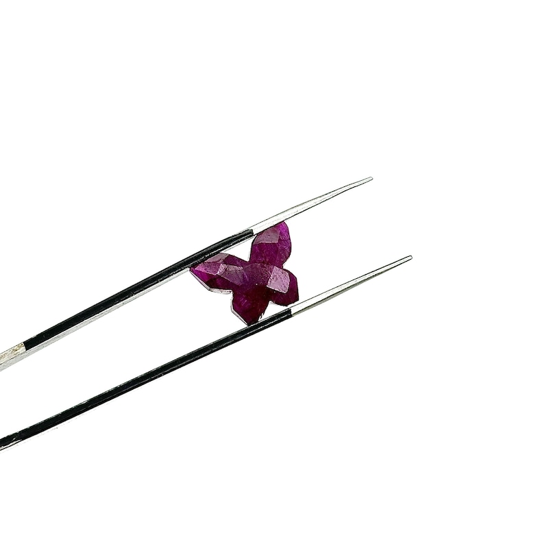 May include: A deep red, faceted gemstone in the shape of a butterfly is held by tweezers. The butterfly gem has multiple facets that reflect light. The tweezers are silver. The gem is likely a ruby.