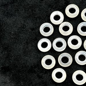 May include: A collection of translucent, off-white, circular beads with a hole in the center, arranged against a dark, textured background. The beads are evenly spaced, creating a pattern. These beads are suitable for jewelry making.