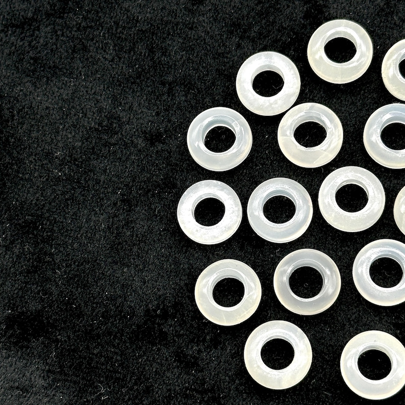 May include: A collection of translucent, off-white, circular beads with a hole in the center, arranged against a dark, textured background. The beads are evenly spaced, creating a pattern. These beads are suitable for jewelry making.