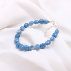 Op de afbeelding: Een lichtblauwe kralenarmband met een zilveren sluiting. De armband heeft ovaalvormige kralen in verschillende blauwtinten. De sieraden worden gepresenteerd op een witte, zijdeachtige stof.