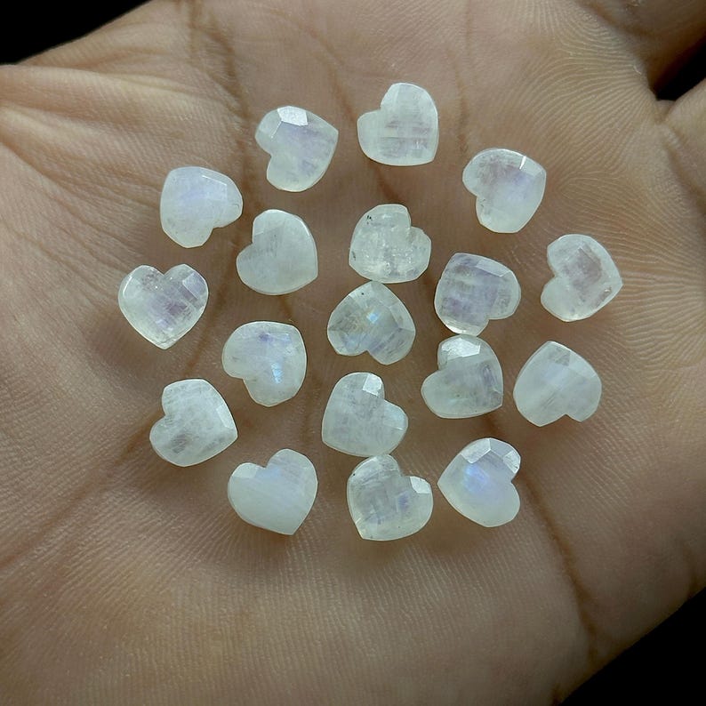 May include: A collection of faceted, heart-shaped moonstone gemstones. The stones are a translucent white with subtle blue and iridescent flashes. The hearts are small and appear to be hand-cut, with multiple facets catching the light.