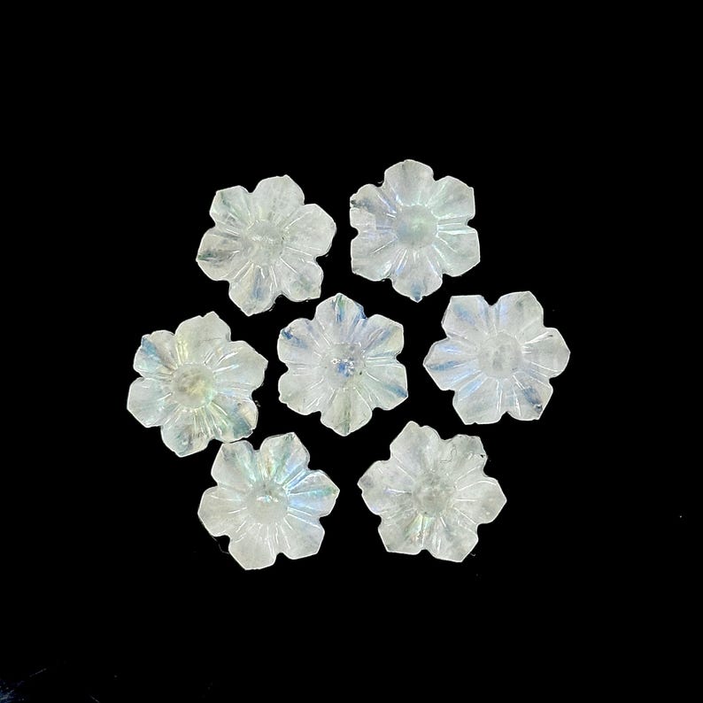 May include: Seven translucent, flower-shaped beads with iridescent qualities are arranged against a black background. Each bead has six petals and a central depression, creating a delicate, floral design. The beads appear to be made of a clear, possibly crystalline material.