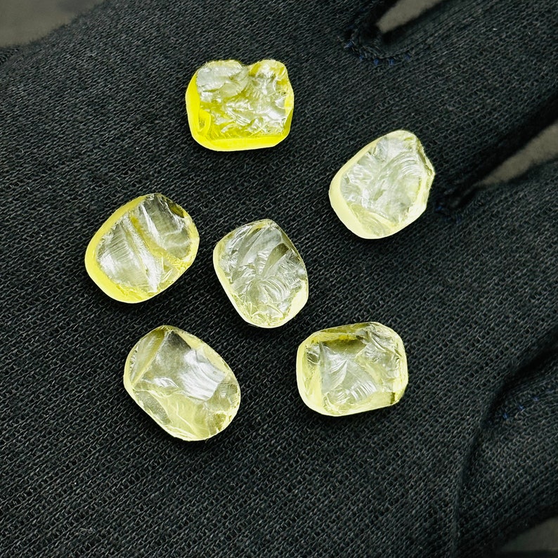 May include: Six yellow faceted gemstones on a black fabric background. The gemstones are irregularly shaped and have a translucent appearance.