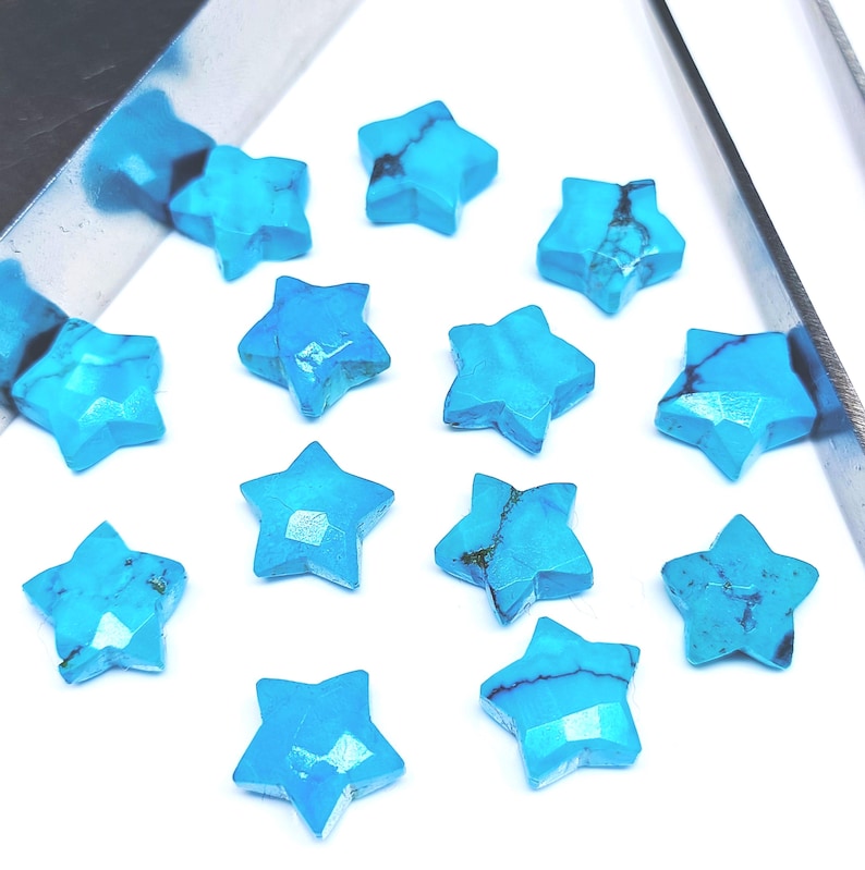 May include: A collection of turquoise star-shaped gemstones. Each star has a faceted surface and a vibrant blue colour with darker veining. The gemstones are arranged on a white surface, with a silver tool partially visible in the upper left corner.