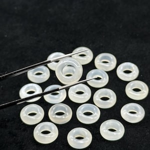 May include: A collection of translucent, ring-shaped beads. The beads are a pale, milky white color and appear to be made of a smooth, polished material. Tweezers are holding one bead, with others scattered on a dark surface.