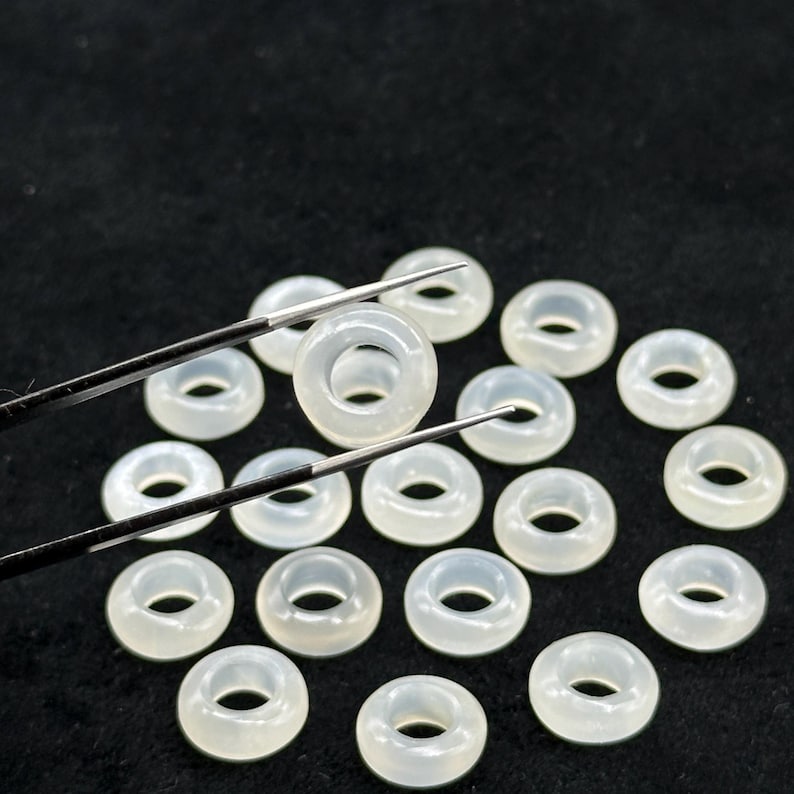 May include: A collection of translucent, ring-shaped beads. The beads are a pale, milky white color and appear to be made of a smooth, polished material. Tweezers are holding one bead, with others scattered on a dark surface.
