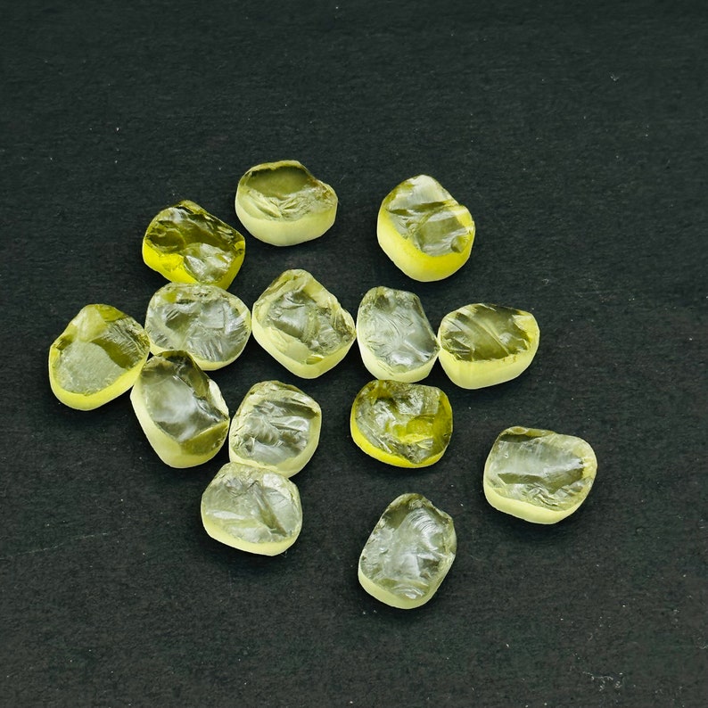 May include: A collection of 13 yellow-green gemstones, some with a slightly cloudy appearance, arranged on a black surface.