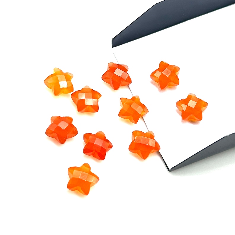 May include: A collection of ten orange star-shaped carnelian gemstones. The stones are arranged on a white surface with a black background.