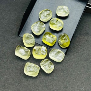 May include: A collection of 12 yellow-green gemstones, some with a more translucent appearance than others, are scattered on a black surface. The gemstones are various shapes and sizes, with some being more rectangular and others more rounded.