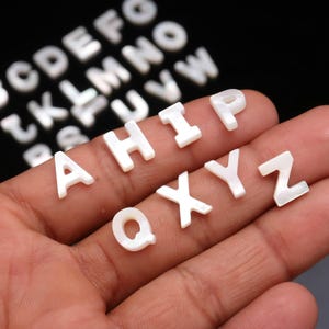 Könnte beinhalten: Eine Sammlung kleiner, weißer, perlmuttartiger Buchstaben. Die Buchstaben sind auf dunklem Hintergrund verstreut, einige werden in einer Hand gehalten. Die Buchstaben sind in einer Serifenschrift und scheinen aus einem muschelartigen Material zu bestehen.