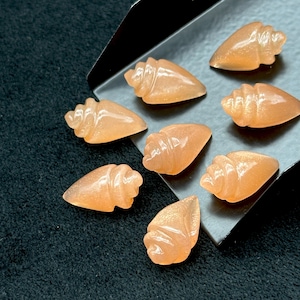 May include: Eight small, carved seashell-shaped stones in shades of peach and pink. The stones are arranged on a grey tray.