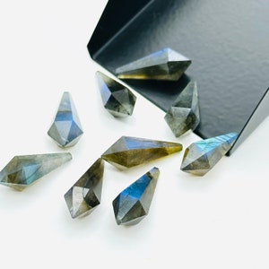 May include: A collection of eight faceted labradorite gemstone points in various shades of grey and blue. The points are arranged on a white surface.
