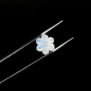 May include: A carved, flower-shaped gemstone with a pearly white hue and subtle blue iridescence. The gem is held by silver tweezers against a black background, highlighting its delicate details and form. The gemstone is likely used for jewelry.