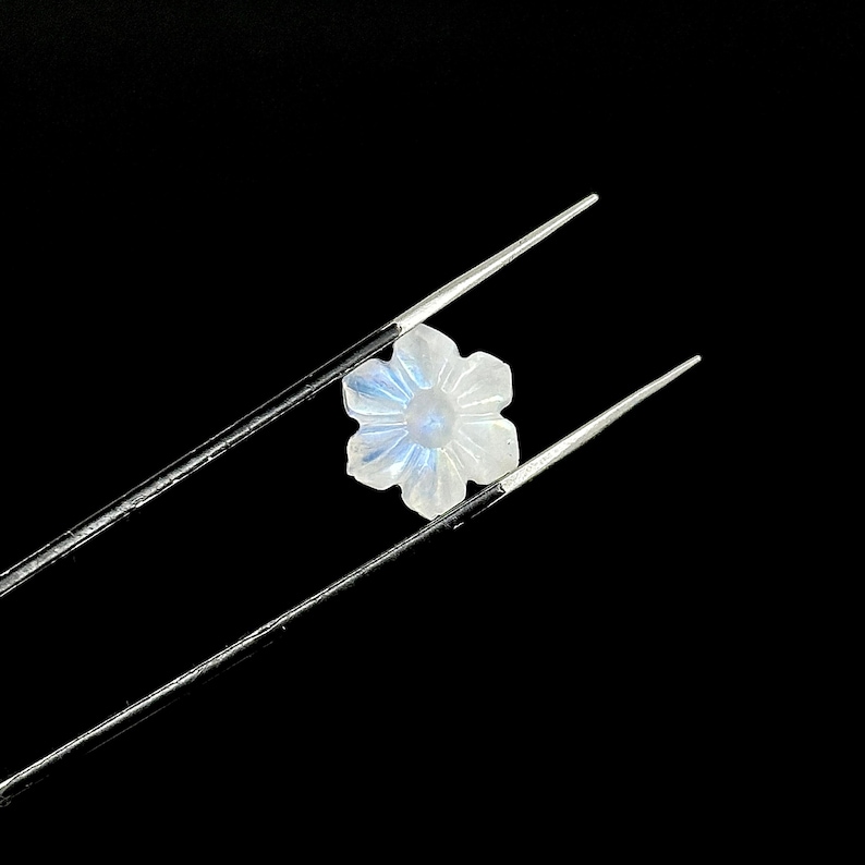 May include: A carved, flower-shaped gemstone with a pearly white hue and subtle blue iridescence. The gem is held by silver tweezers against a black background, highlighting its delicate details and form. The gemstone is likely used for jewelry.