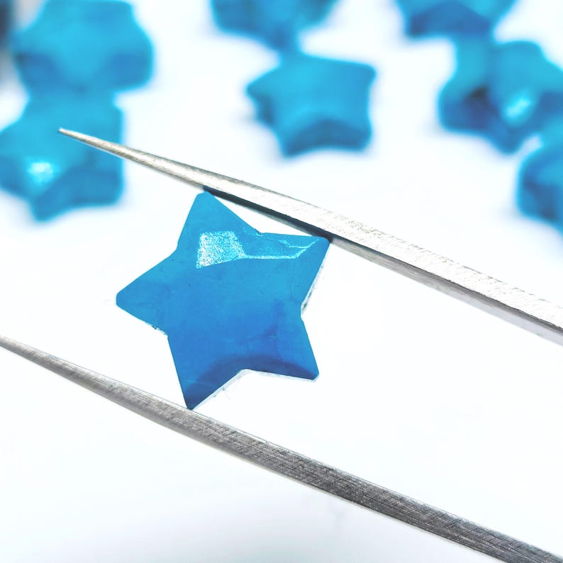 May include: A close-up shot of a vibrant blue star-shaped bead held by silver tweezers. Other blue star beads are visible in the background, set against a white background. The beads appear to be made of a solid material.