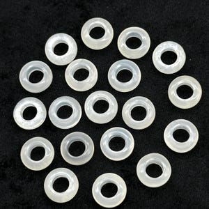May include: A collection of translucent, off-white, donut-shaped beads. Each bead has a central hole and a smooth, rounded surface. The beads are scattered on a dark background, creating a contrast that highlights their shape and color. These beads are suitable for jewelry making.