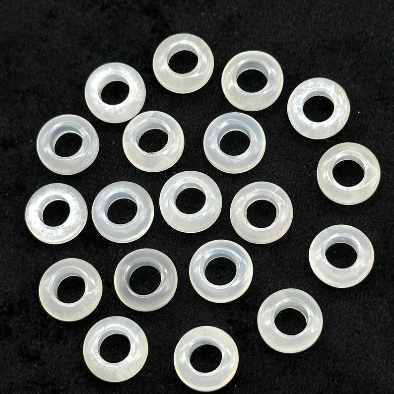 May include: A collection of translucent, off-white, donut-shaped beads. Each bead has a central hole and a smooth, rounded surface. The beads are scattered on a dark background, creating a contrast that highlights their shape and color. These beads are suitable for jewelry making.