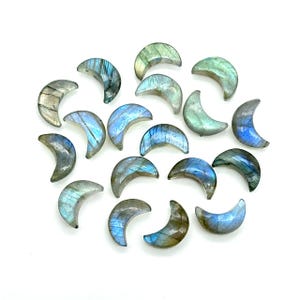 May include: A collection of polished labradorite crescent moon gemstones in various shades of blue, green, and gray. The gemstones are arranged on a white surface, showcasing their iridescent qualities and unique patterns.