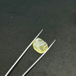 May include: A yellow gemstone with a striated pattern is held by a pair of tweezers against a dark gray background.