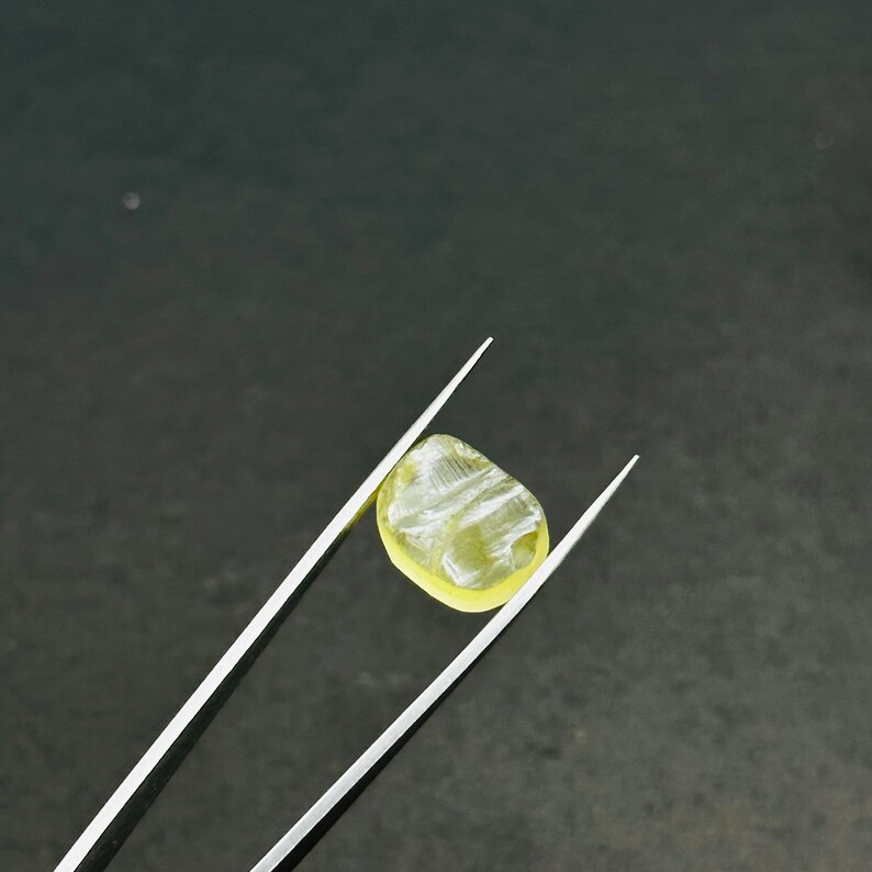May include: A yellow gemstone with a striated pattern is held by a pair of tweezers against a dark gray background.