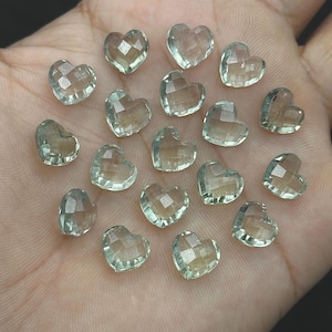 May include: A collection of faceted, heart-shaped gemstones in a pale green hue. The gemstones are arranged in a hand, showcasing their clarity and multiple facets. These heart-shaped stones are ideal for jewelry making or crafting.