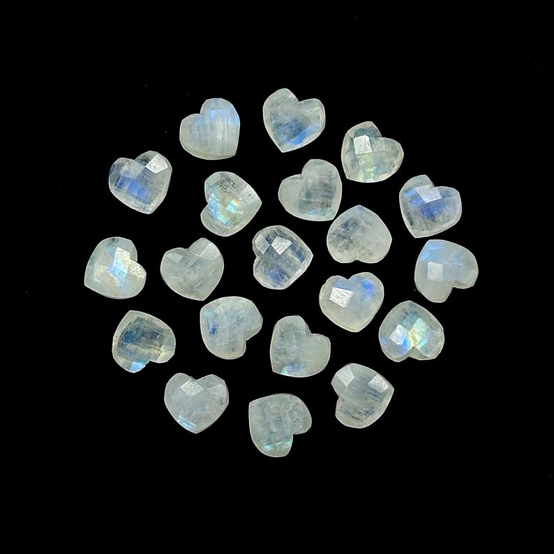 May include: A collection of heart-shaped moonstone gemstones. Each stone has a faceted surface, displaying a range of colors including white, blue, and iridescent flashes. The gemstones are arranged against a black background.