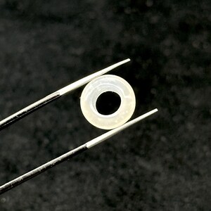 May include: A small, translucent, circular object with a dark center is held by a pair of tweezers. The object appears to be a ring or a washer, possibly made of a light-colored material. The background is a dark, solid color.