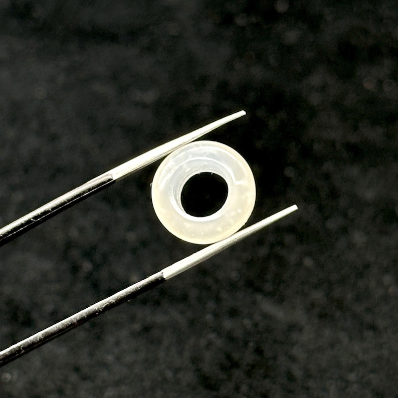 May include: A small, translucent, circular object with a dark center is held by a pair of tweezers. The object appears to be a ring or a washer, possibly made of a light-colored material. The background is a dark, solid color.