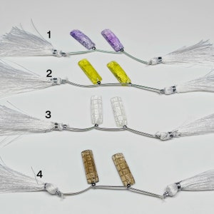 May include: Four sets of two decorative glass beads with white tassels. The beads are in different colors and patterns: purple, yellow, white, and brown.