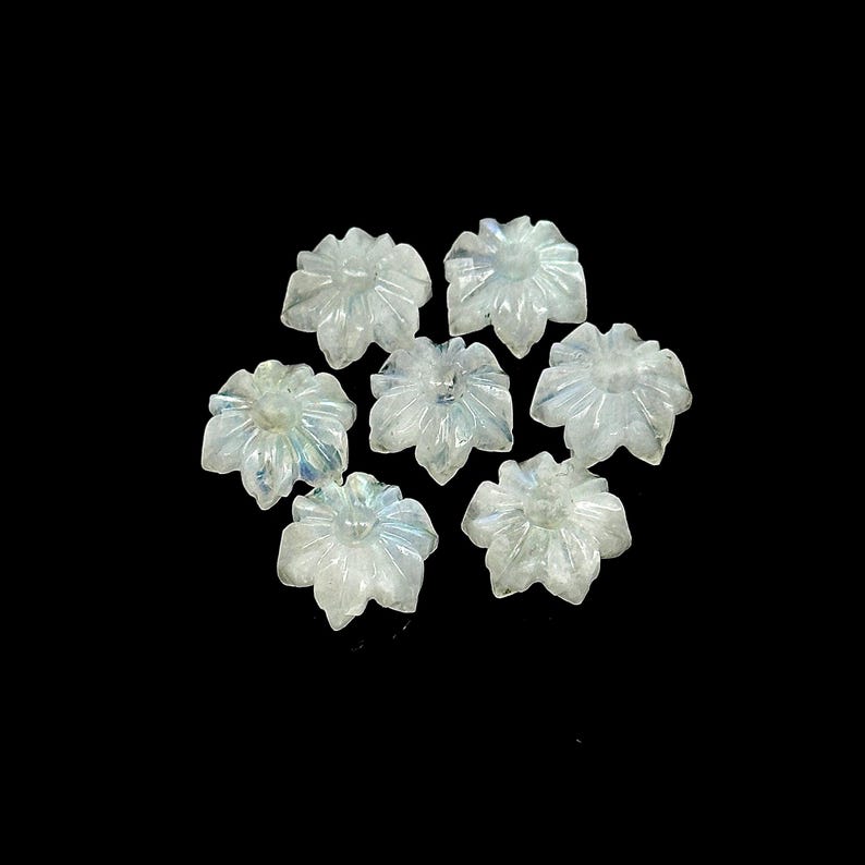 May include: Seven carved, translucent flower-shaped gemstones with a pearlescent sheen. The petals are detailed with carved lines, and the gems are arranged against a black background. These gemstones could be used for jewelry making or other crafts.