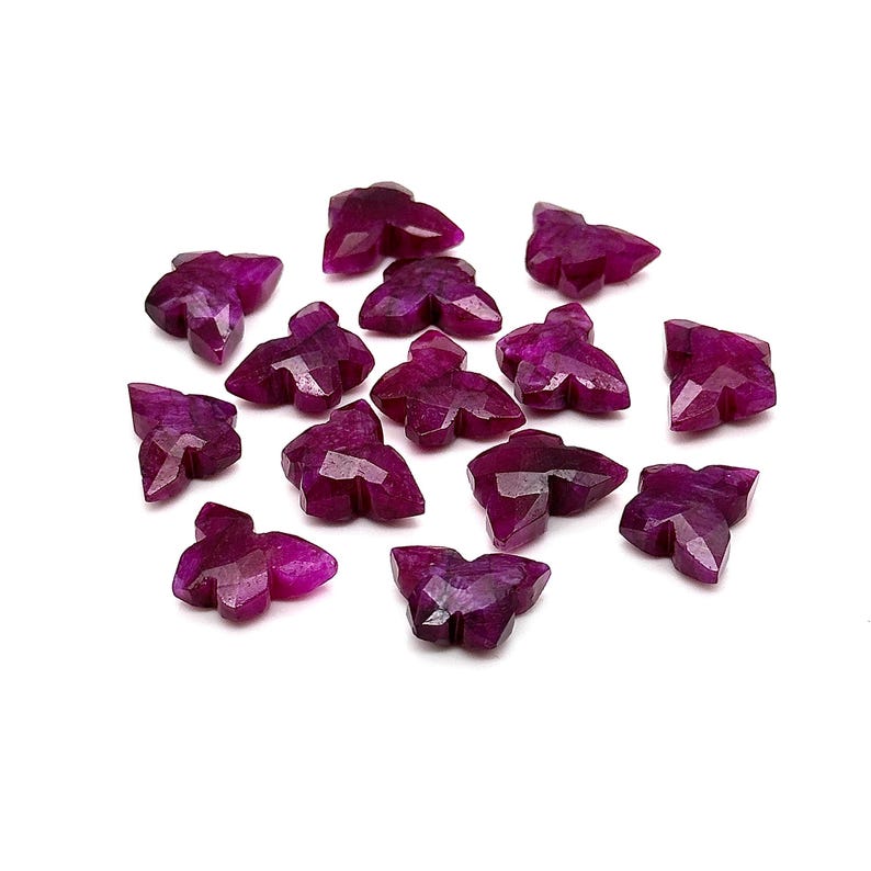 May include: A scattering of faceted, deep magenta gemstone beads, each carved in a stylized leaf shape. The beads are displayed on a white surface, highlighting their geometric cuts and rich colour. Suitable for jewellery making or crafting.