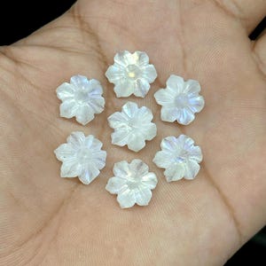 May include: Seven iridescent, carved flower-shaped gemstones. The gemstones are white with subtle blue and purple hues. The flowers have detailed petal carvings and are arranged in a circular pattern, ideal for jewelry making or crafts.