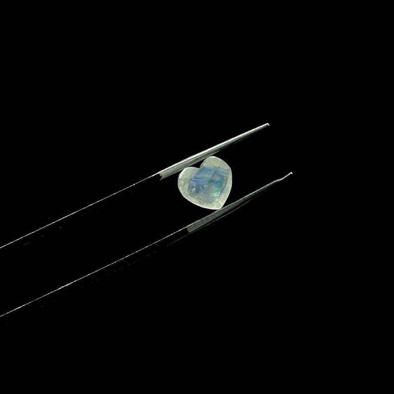 May include: A heart-shaped, iridescent gemstone is held by a pair of tweezers against a black background. The gem displays a mix of pale blue, white, and subtle rainbow hues. The tweezers are silver and angled.