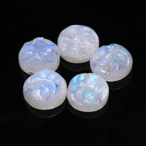 Può includere: Cinque gemme rotonde di pietra di luna scolpite. Ogni pietra presenta un'incisione di un volto e mostra sfumature iridescenti blu e bianche. Le gemme sono disposte su una superficie nera.