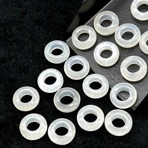 May include: A collection of translucent, ring-shaped beads. The beads are a pale, milky white color and have a smooth, polished surface. They are arranged on a dark, textured surface, with some beads partially overlapping.