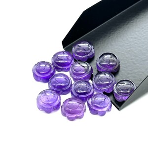 May include: A group of 12 purple amethyst cabochons with a carved smiley face design. The cabochons are scattered on a white surface, with some spilling out of a black tray.