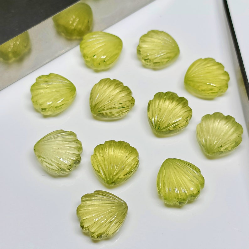 May include: A collection of eleven green gemstone cabochons carved in the shape of seashells. The cabochons are arranged on a white surface.