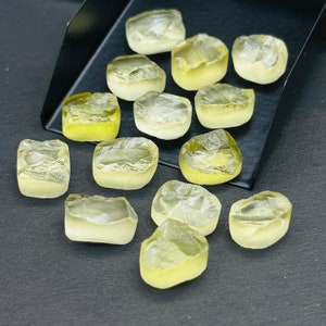 May include: A collection of 14 faceted, translucent yellow gemstones, likely citrine, displayed on a black tray. The stones are various sizes and shapes, with some having a more rectangular shape and others more rounded.