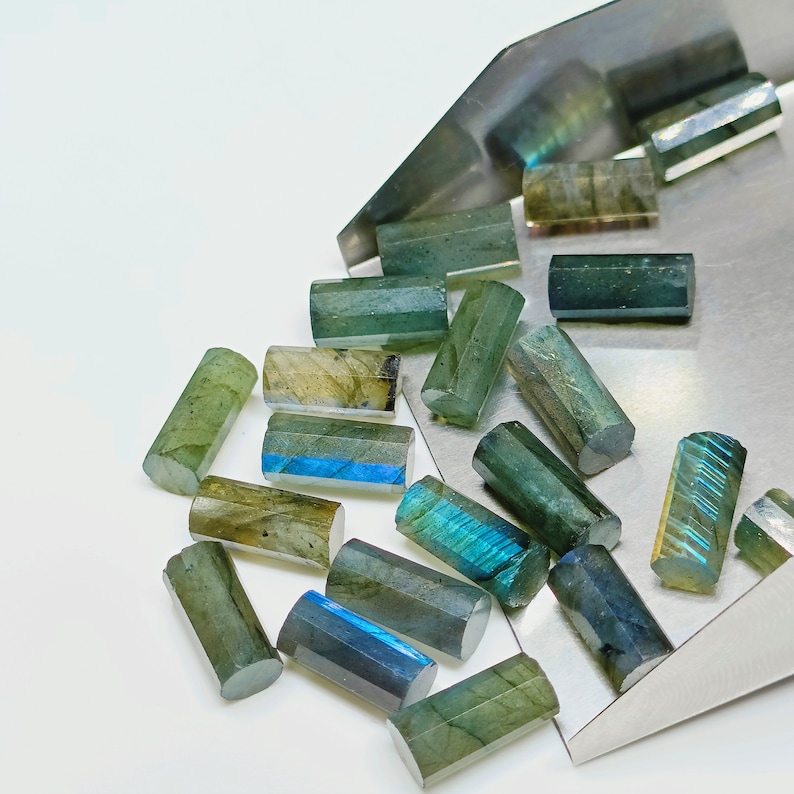 May include: A collection of green and blue labradorite gemstone beads in a variety of shapes and sizes. The beads are arranged in a pile on a metal surface.