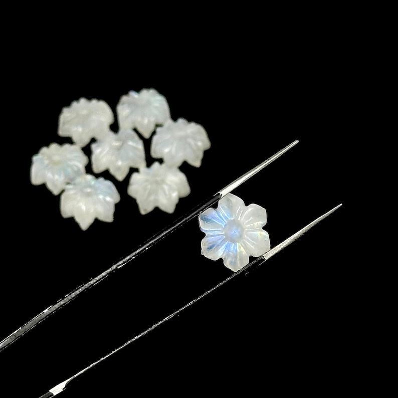 May include: A collection of small, white, flower-shaped gemstones. One gemstone is held by tweezers, showcasing its delicate petals and iridescent blue and white hues. The other gemstones are scattered on a black background.