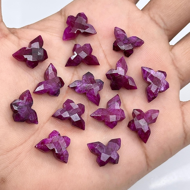 May include: A selection of magenta-coloured, butterfly-shaped gemstones. Each gemstone is multi-faceted, creating a sparkling effect. The butterfly shapes are consistent in size and design, with a faceted cut.