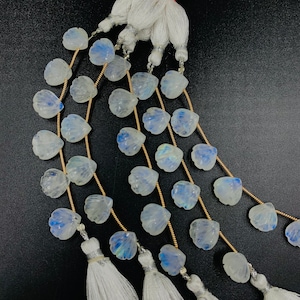 Op de afbeelding: Een streng witte maansteen kralen, gesneden in een schelpvorm, met een regenboogkleurige, iriserende glans. De kralen zijn geregen op een gouden draad en hebben witte kwastjes aan de uiteinden.