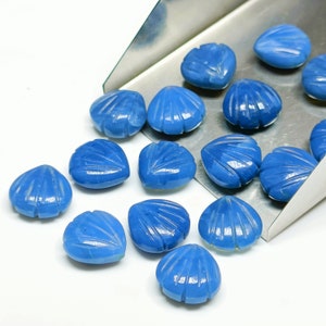 Puede incluir: Un grupo de 14 cabujones de corazón de piedra preciosa azul con un diseño de concha tallada. Las piedras preciosas están dispersas sobre una superficie blanca y una pala de plata.