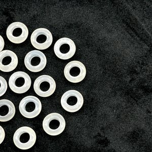 May include: A collection of translucent, ring-shaped beads with a central hole. The beads are arranged on a black surface, creating a contrast between the light color of the beads and the dark background. The beads appear to be made of a smooth, possibly polished material.