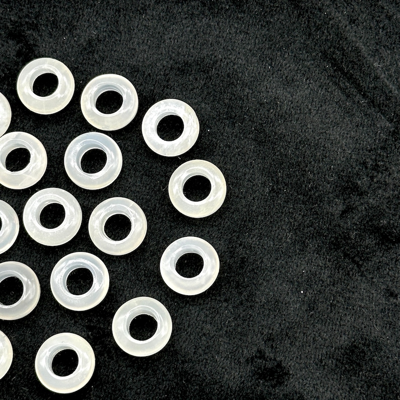 May include: A collection of translucent, ring-shaped beads with a central hole. The beads are arranged on a black surface, creating a contrast between the light color of the beads and the dark background. The beads appear to be made of a smooth, possibly polished material.