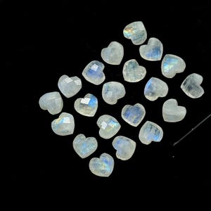 May include: A collection of heart-shaped moonstone gemstones. The stones are a translucent white with iridescent blue and yellow flashes. The faceted surfaces catch the light, creating a sparkling effect. The gemstones are arranged on a black background.